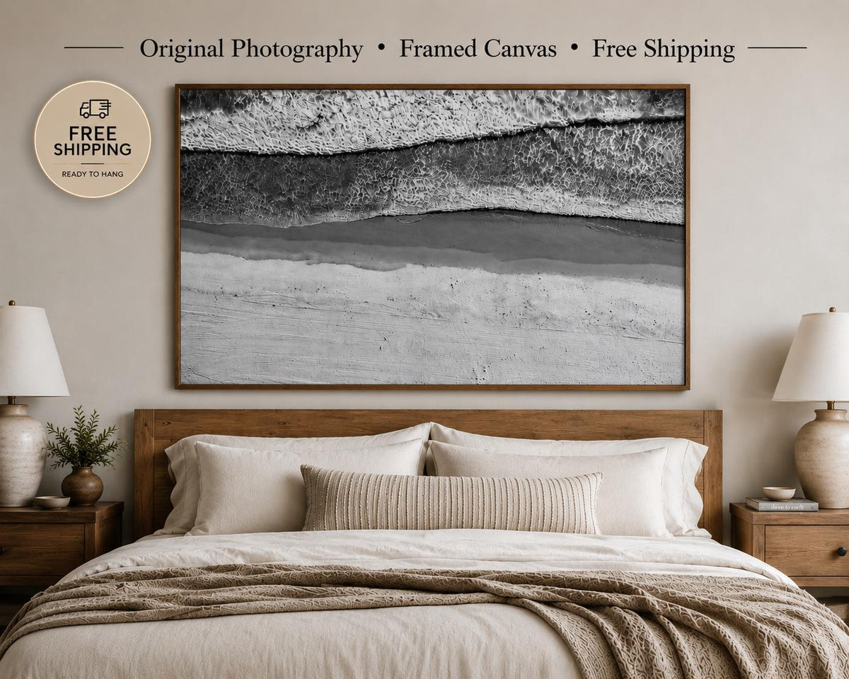 Original black and white aerial beach photography printed on premium framed canvas, displayed above a bed in a modern minimalist bedroom. Available as a single horizontal canvas or triptych wall art with free shipping.
