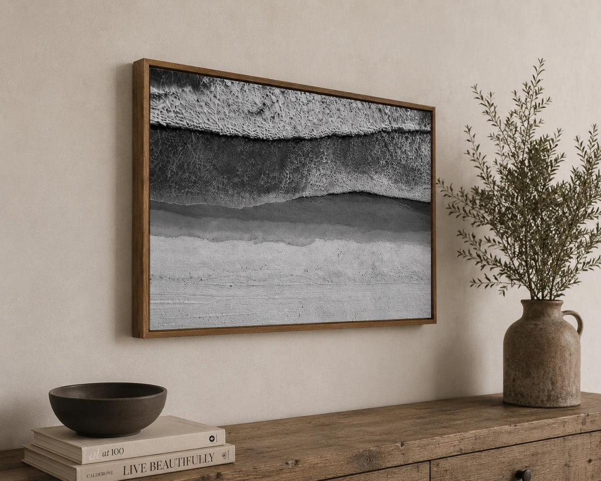 May include: A rectangular wall art piece featuring a black and white aerial photograph of ocean waves crashing onto a sandy beach, enclosed in a thin wooden frame. The image displays textured white foam, dark water, and a light-colored shoreline.