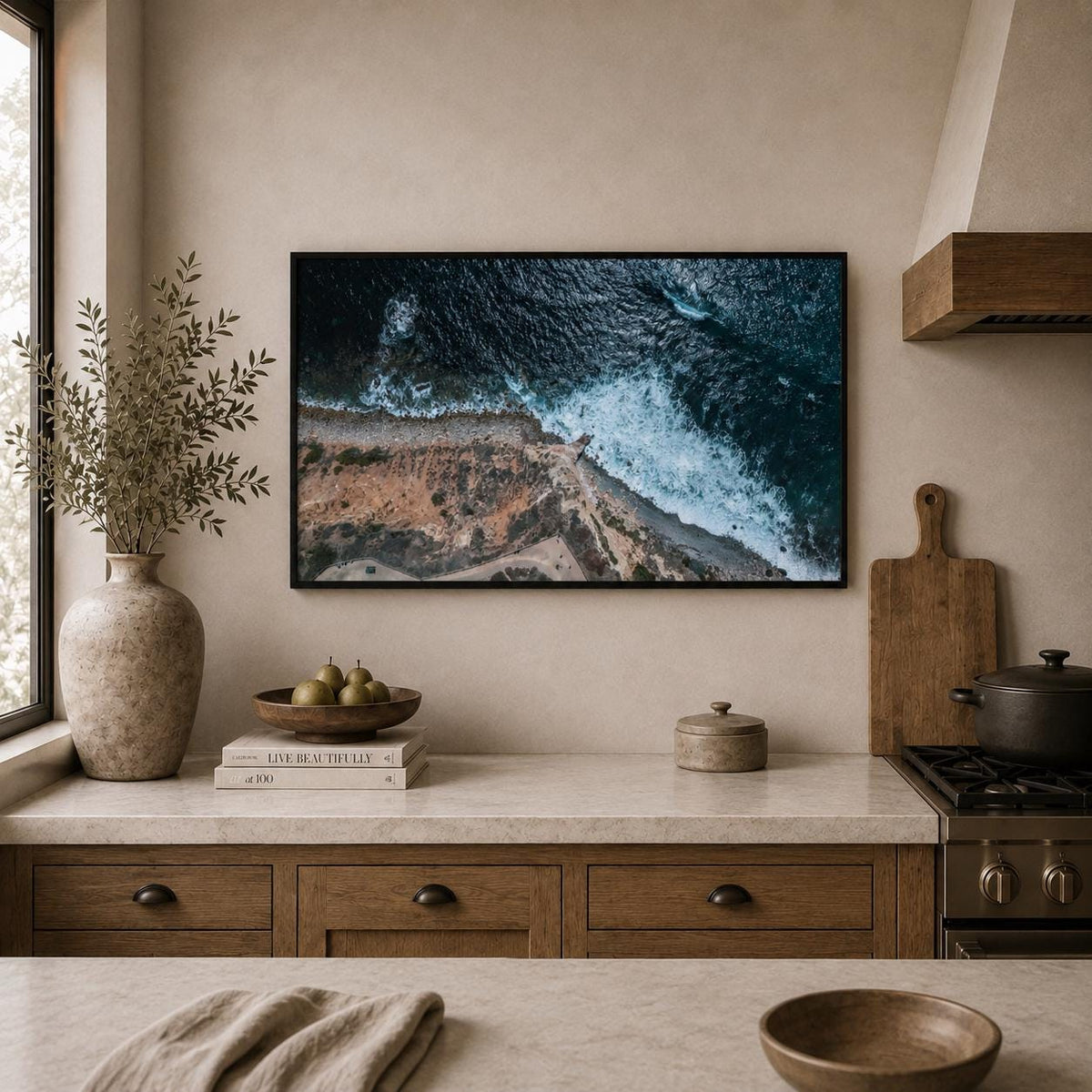 May include: A rectangular framed wall art print featuring an aerial view of dark blue ocean waves crashing against a rocky, tan-colored coastline. The artwork is displayed on a beige wall above a kitchen counter with a vase, books, and a bowl.