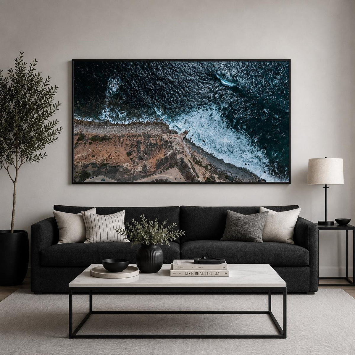 May include: A large rectangular wall art print featuring an aerial view of a dark blue ocean meeting a rocky brown coastline with white crashing waves, framed in black and mounted above a dark gray sofa with light gray and striped throw pillows.