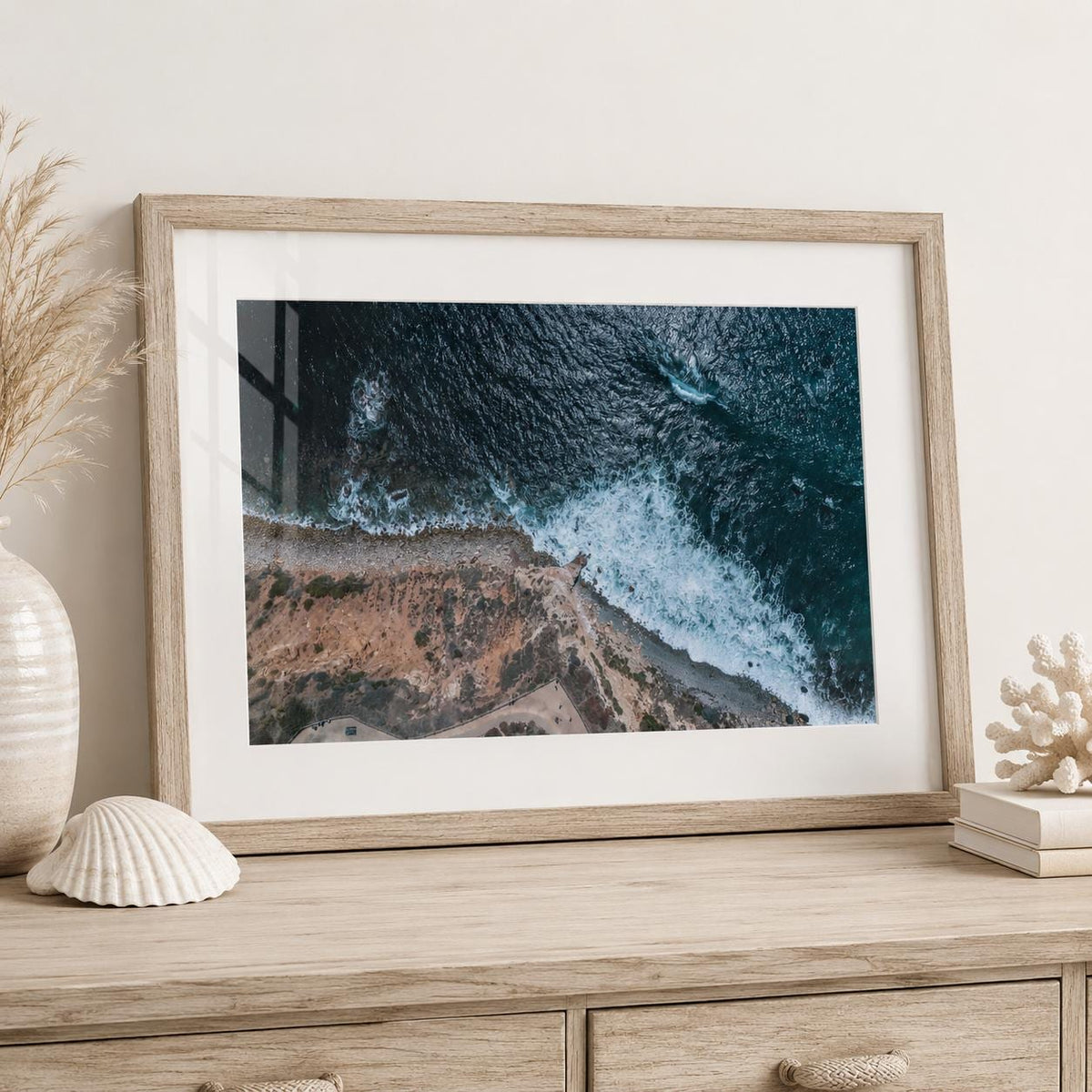 May include: A framed wall art print featuring an aerial view of a rugged coastline where dark blue ocean waves crash against a rocky shore. The print is housed in a light-toned wooden frame with a white mat, displaying a high-contrast coastal scene.