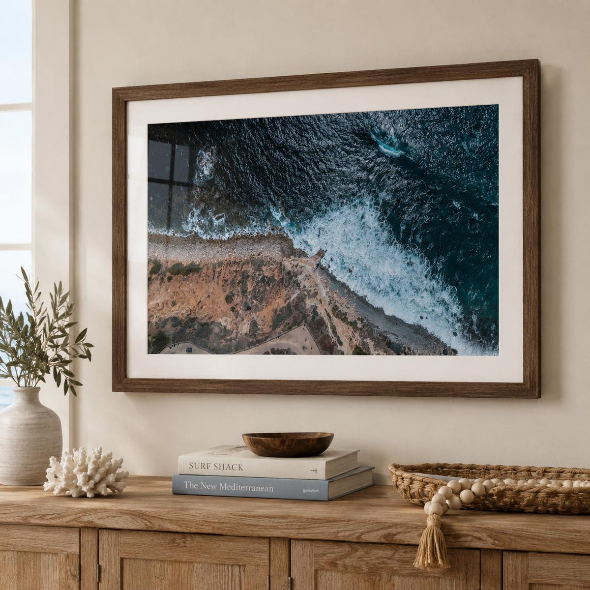 May include: A framed wall art print featuring an aerial view of a rugged coastline where dark blue ocean waves crash against a rocky, tan-colored shore. The print is enclosed in a dark brown wooden frame with a wide white mat border.
