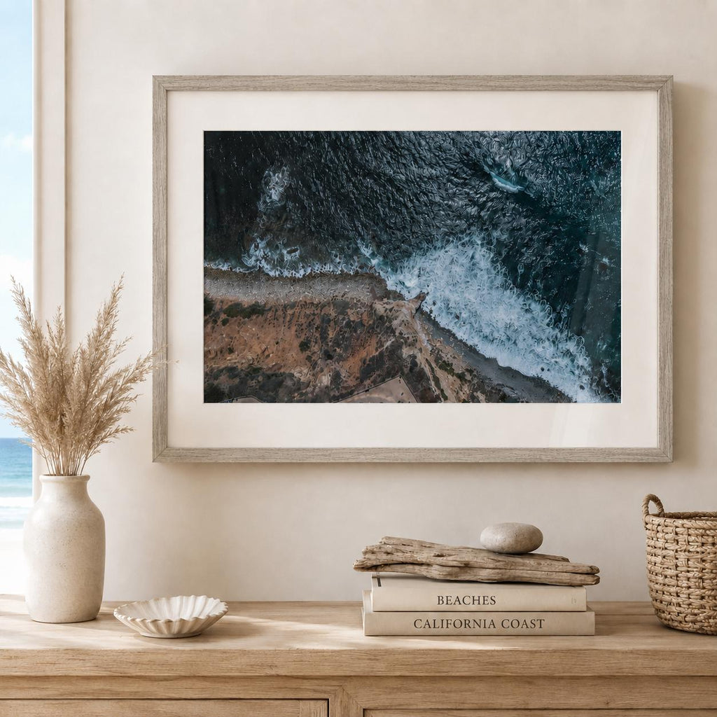 May include: A framed landscape photograph showing an aerial view of dark blue ocean waves crashing against a rocky brown coastline. The print is displayed in a light wood frame with a white mat, positioned above a stack of books and a vase.