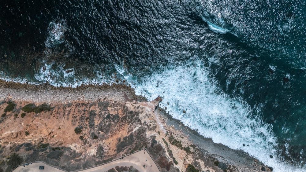 May include: An aerial view of a rugged coastline where dark blue ocean waves crash against a rocky shore. The scene features a transition from deep water with white foam to a pebbled beach and a dry, tan-colored cliffside with sparse vegetation.