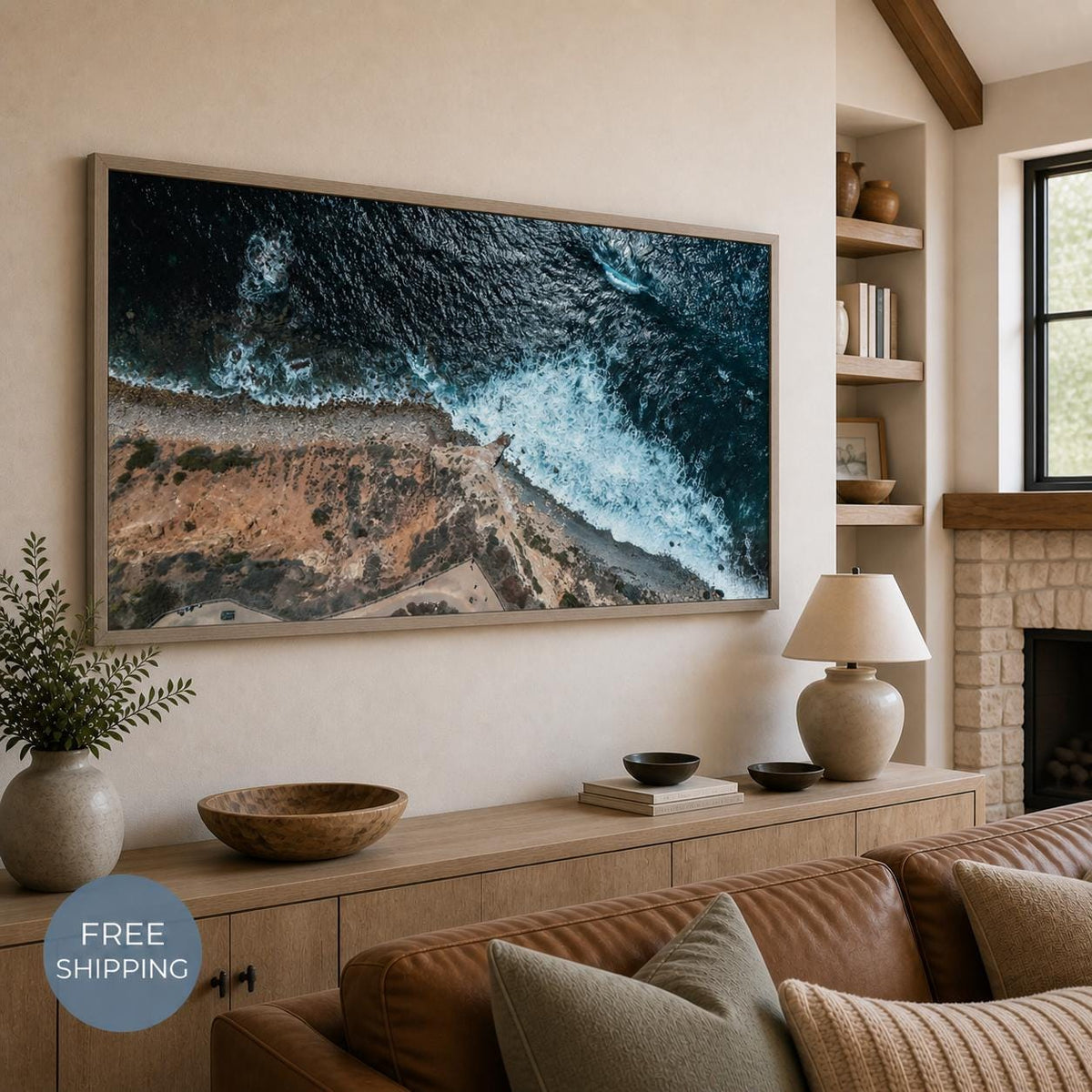 May include: A rectangular wall art print featuring an aerial view of a rugged coastline where dark blue ocean waves crash against a rocky, tan-colored shore. The print is framed in a thin, light-toned wood frame and mounted on a beige wall.