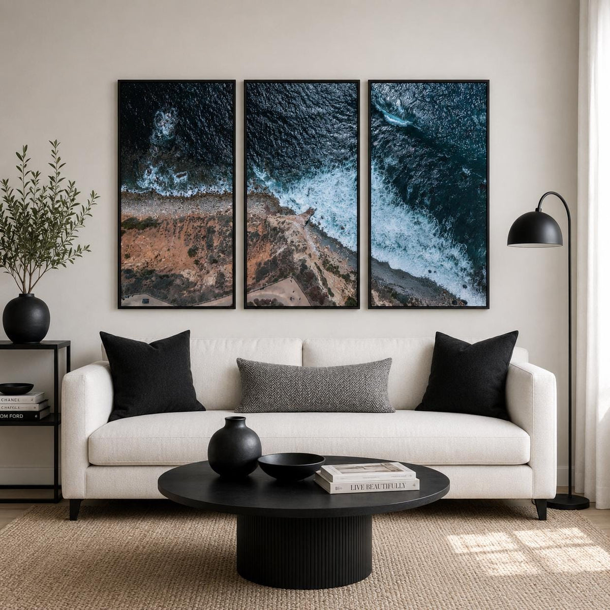 May include: A triptych wall art set featuring an aerial view of a rugged coastline where dark blue ocean waves crash against rocky cliffs. The three vertical framed prints are displayed above a white sofa with black pillows and a round black coffee table.