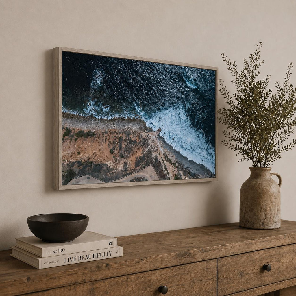 May include: A rectangular framed wall art print featuring an aerial view of a rugged coastline where dark blue ocean waves crash against a sandy shore. The print is displayed in a light wood frame and hangs on a neutral-colored wall.