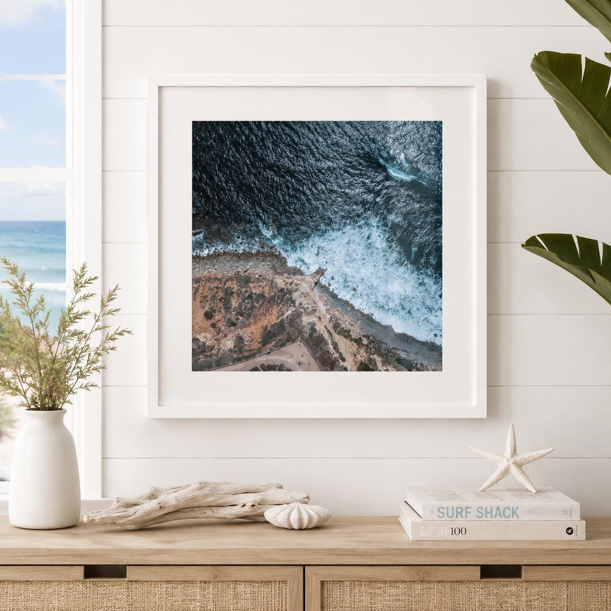 May include: A square framed wall art print featuring an aerial view of a rugged coastline where dark blue ocean waves crash against a rocky, tan-colored shore. The print is displayed in a white frame, capturing the contrast between the sea and land.