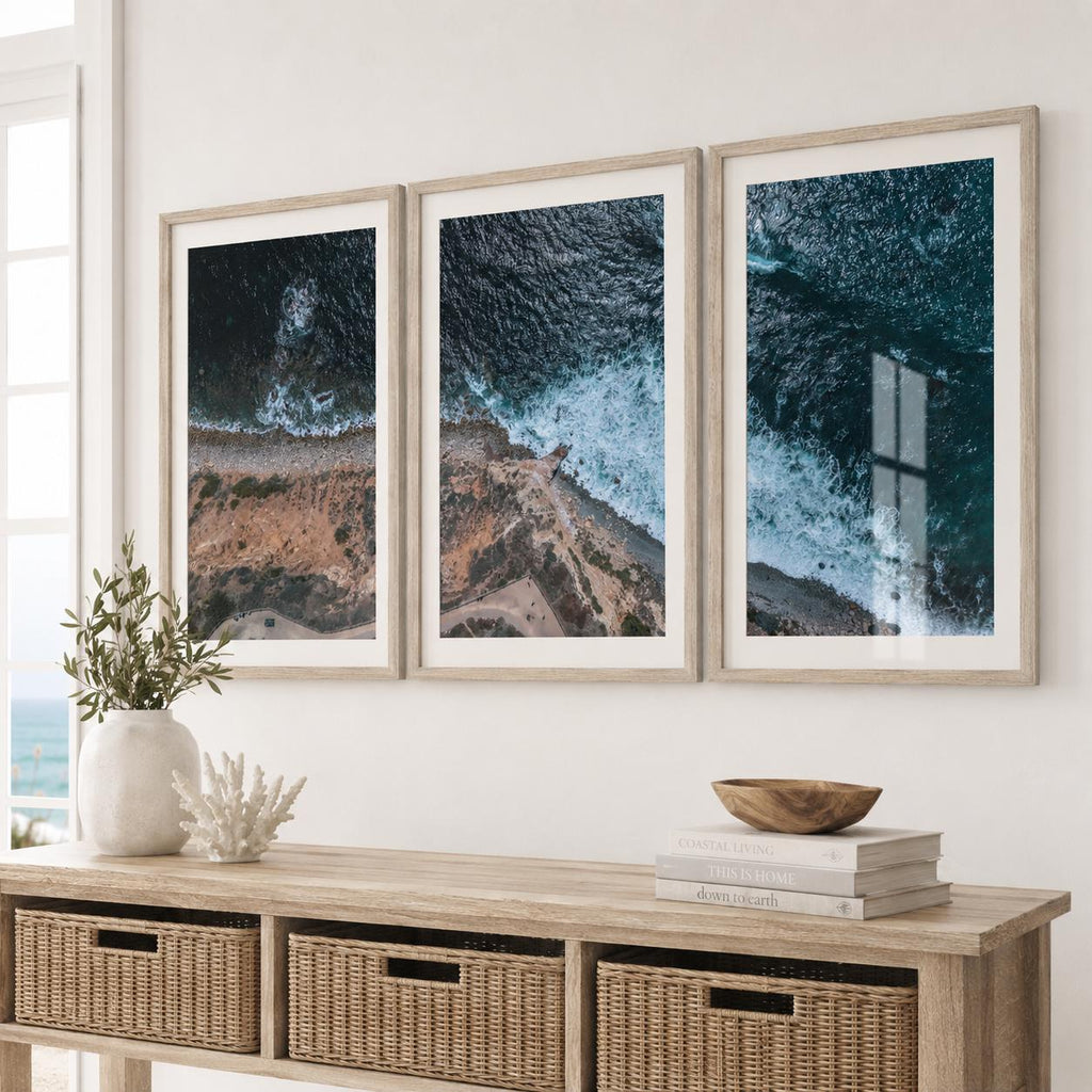 May include: A triptych of framed wall art prints featuring aerial photography of a rugged coastline where dark blue ocean waves meet a rocky shore. The three vertical prints are displayed in light wood frames, showcasing the contrast between the deep sea and the textured earth.