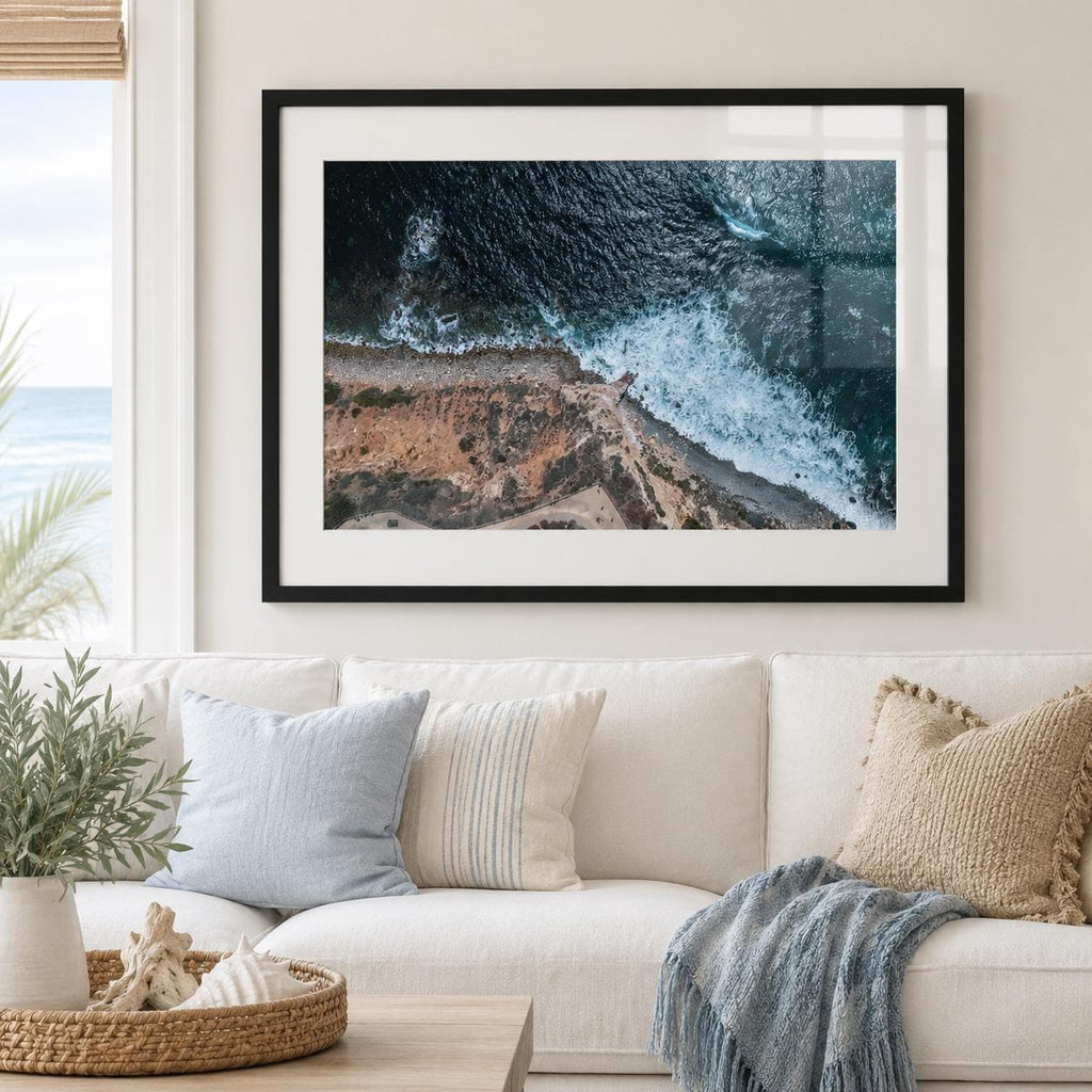 May include: A framed landscape photograph featuring an aerial view of a rugged coastline where dark blue ocean waves crash against a rocky, tan-colored shore. The print is encased in a simple black frame with a wide white mat border.