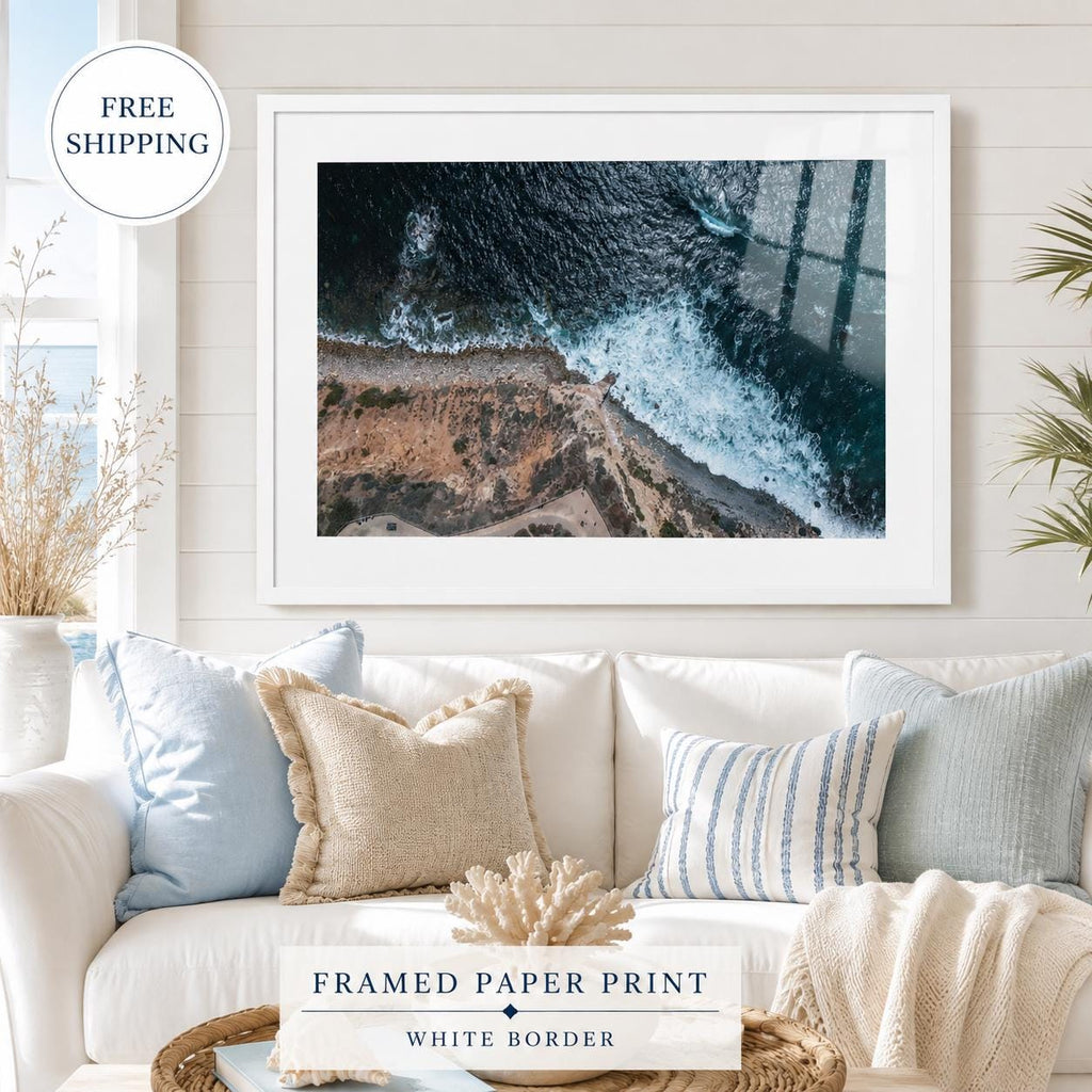 Framed aerial ocean photography print with a white mat and white frame, featuring deep blue waves crashing along a rocky coastline, displayed in a bright coastal-style interior with subtle glass reflection.