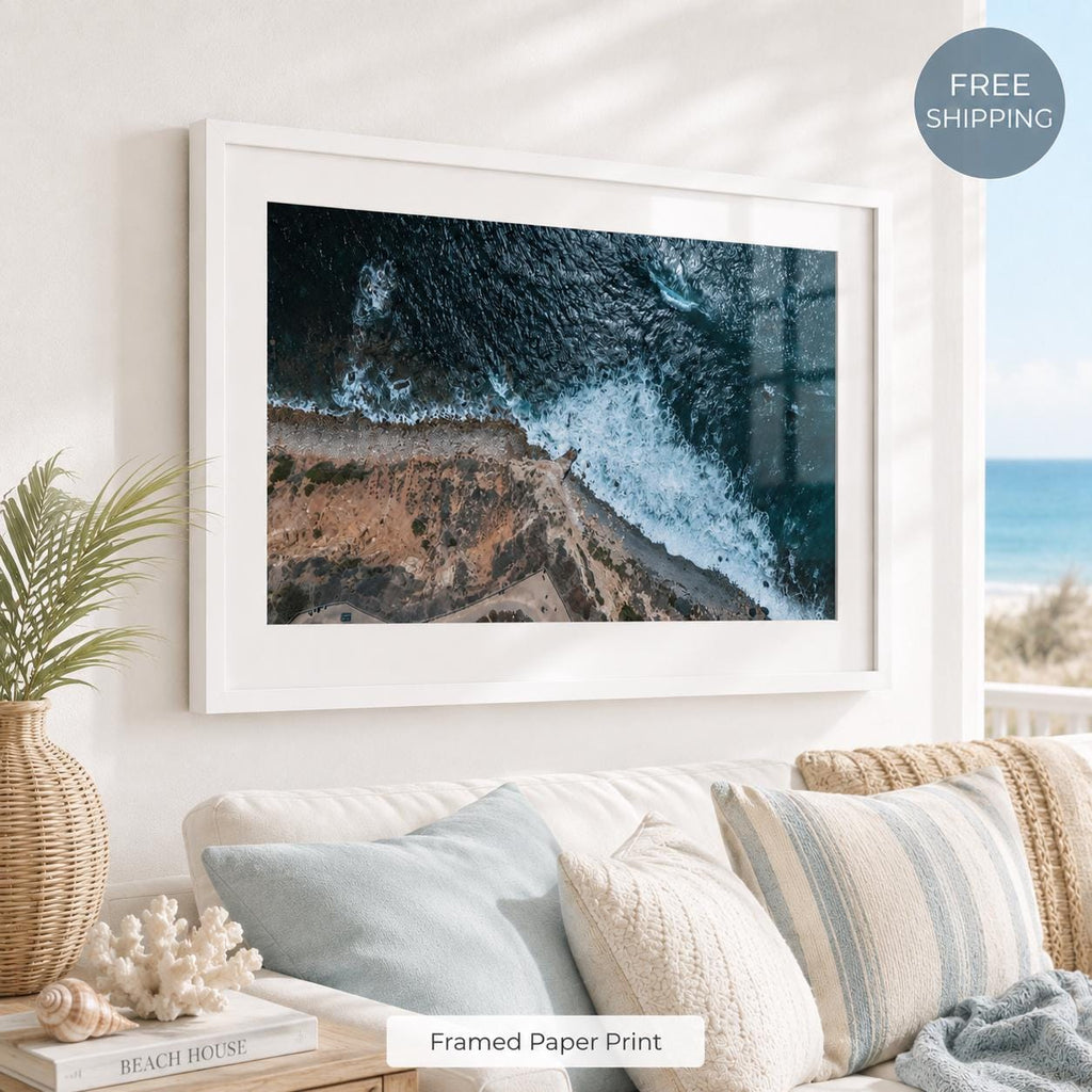 Framed aerial ocean photography print with a white mat and white frame, showing deep blue waves crashing along a rocky coastline in a bright beach-inspired living room with soft blue pillows and coastal decor.