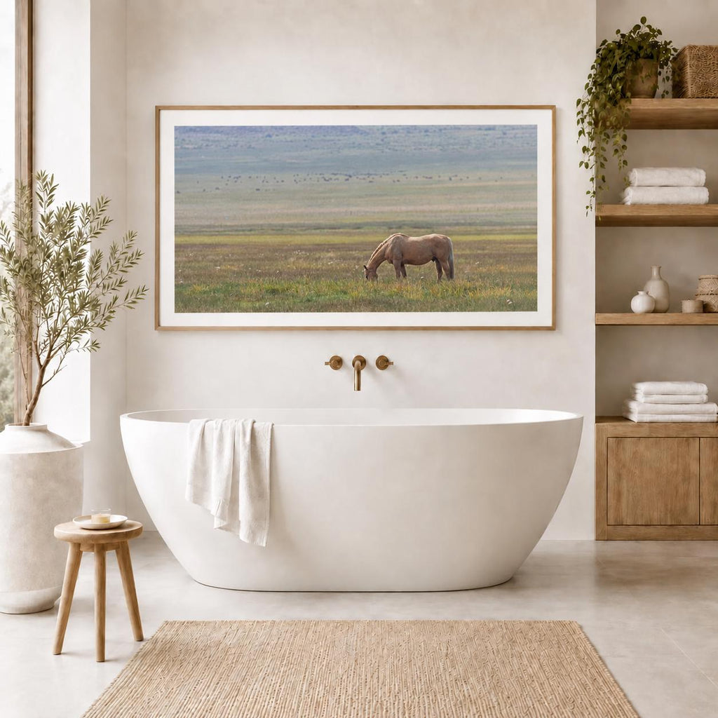 May include: A rectangular framed landscape print featuring a brown horse grazing in a vast, grassy field. The artwork is mounted on a light-colored wall above a white freestanding oval bathtub with a white towel draped over the side.