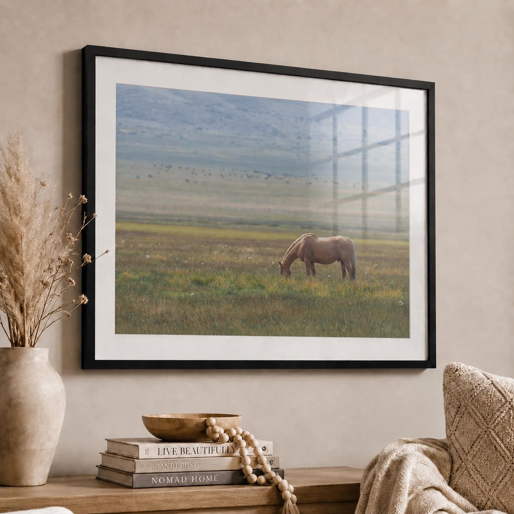Side-angle view of a grazing horse photography print in a black frame with white mat and glossy acrylic glazing, showing realistic window reflections above a neutral wood console.