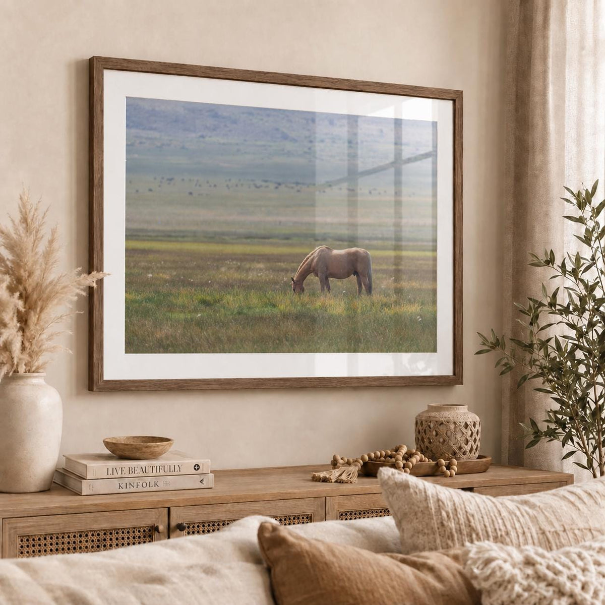 May include: A framed landscape wall art print featuring a light brown horse grazing in a vast, grassy field under a soft, overcast sky. The artwork is enclosed in a simple wooden frame with a white mat, displayed on a wall above a wooden console.
