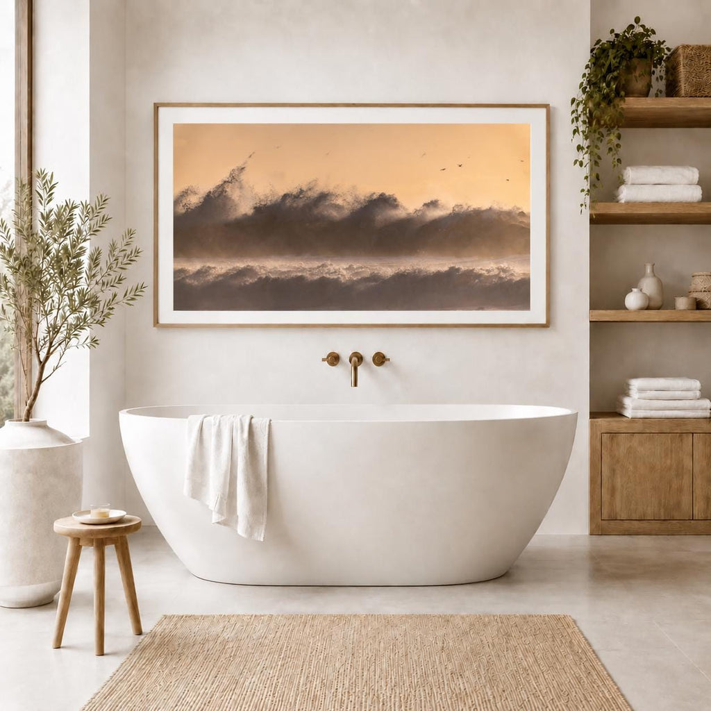 Golden-hour crashing ocean wave photography in a natural wood frame with a white mat above a freestanding bathtub in a bright neutral bathroom with plants and wood shelving.