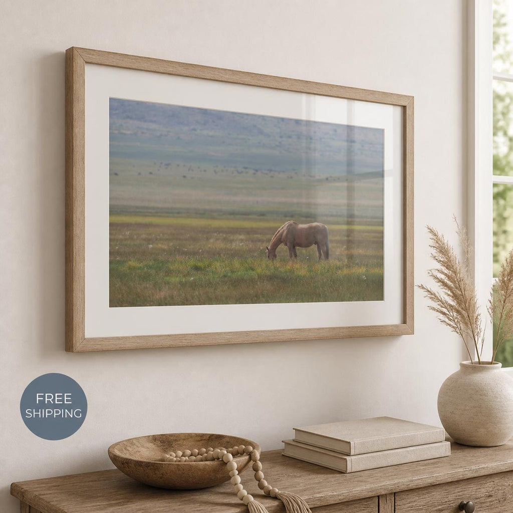 May include: A framed landscape photograph featuring a light brown horse grazing in a vast, grassy field under a soft, overcast sky. The artwork is enclosed in a light-toned wooden frame with a wide white mat, displayed on a neutral-colored wall.