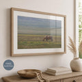 May include: A framed landscape photograph featuring a light brown horse grazing in a vast, grassy field under a soft, overcast sky. The artwork is enclosed in a light-toned wooden frame with a wide white mat, displayed on a neutral-colored wall.