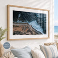 May include: A rectangular wall art piece featuring an aerial view of a rugged coastline where dark blue ocean waves crash against a sandy beach and rocky cliffs, enclosed in a light-toned wooden frame with a wide white mat border.
