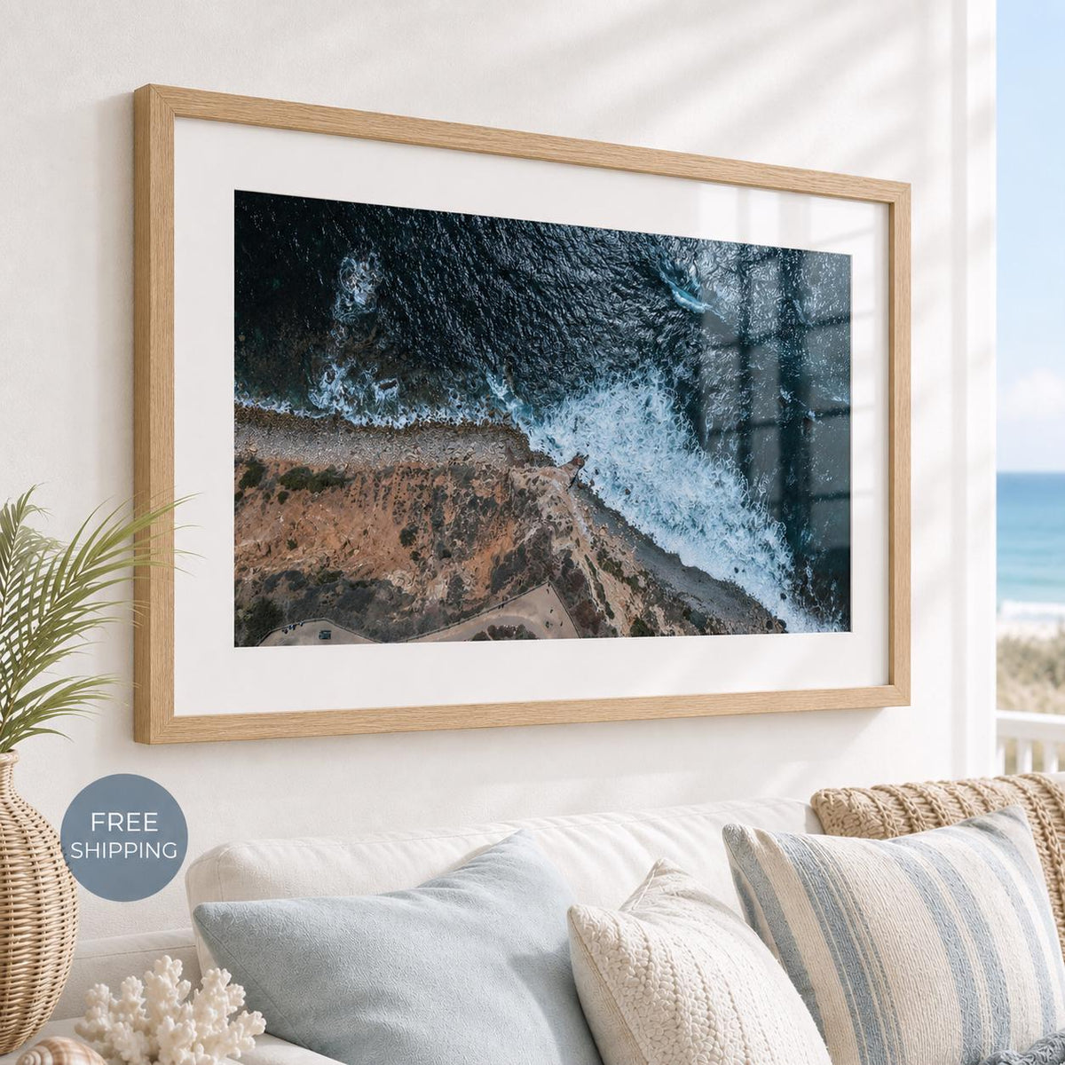 May include: A rectangular wall art piece featuring an aerial view of a rugged coastline where dark blue ocean waves crash against a sandy beach and rocky cliffs, enclosed in a light-toned wooden frame with a wide white mat border.