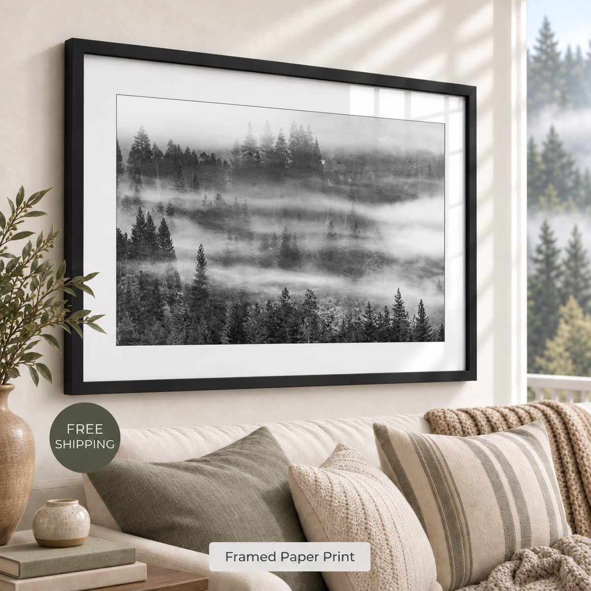 Black-and-white photograph of evergreen forest hills layered with drifting fog, shown with a white mat and black frame above a neutral sofa. Room mockup includes a free-shipping badge.