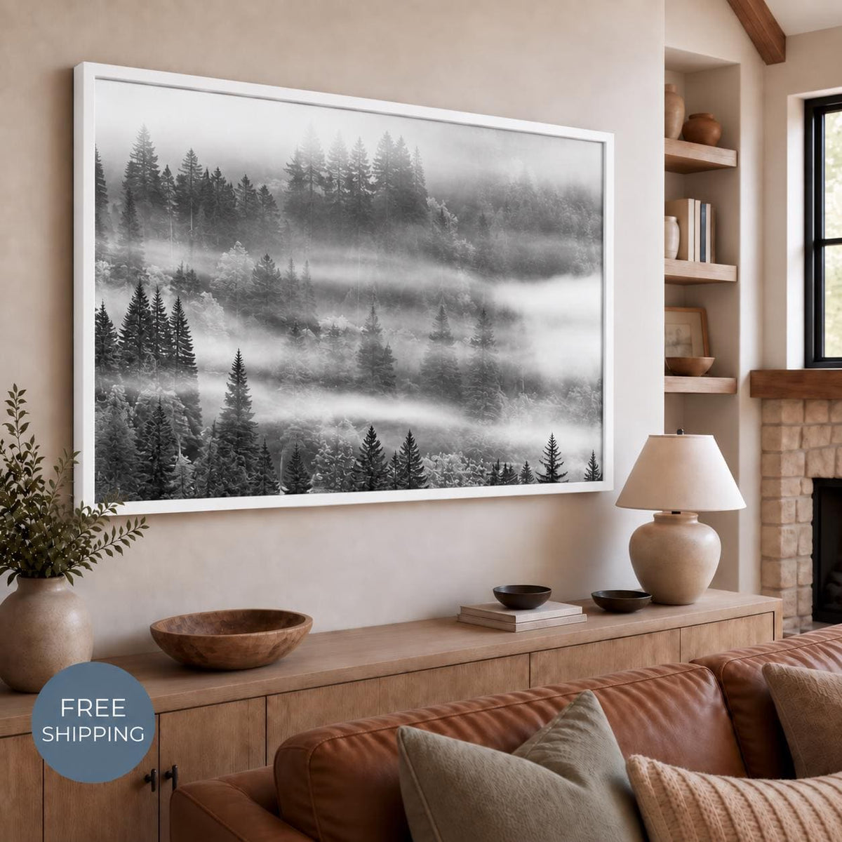 Black and white misty forest canvas in a white floating frame above a wooden console and brown sofa, with evergreen trees fading into horizontal bands of fog. A badge reads Free Shipping.