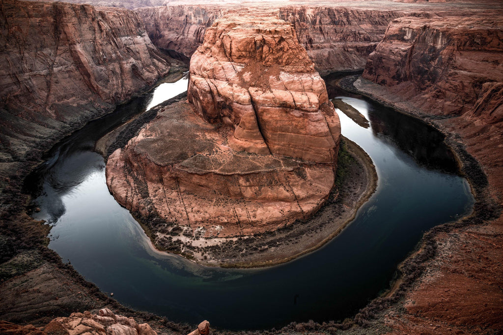 Original Horseshoe Bend landscape photograph near Page, Arizona. The blue-green Colorado River curves around a towering rust-colored sandstone formation, with layered canyon walls and rocky foreground visible across the scene.